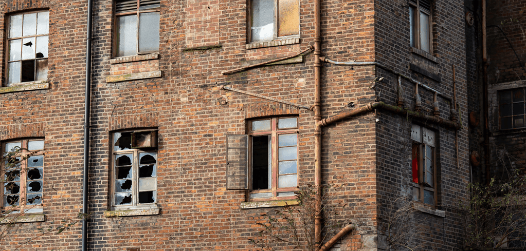 Abandoned brick building with broken windows, exposed pipes, and deteriorating facade showing signs of urban decay.