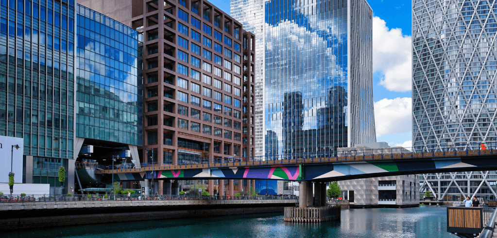 Modern glass skyscrapers in urban financial district with colorful bridge spanning a canal under blue sky with clouds.