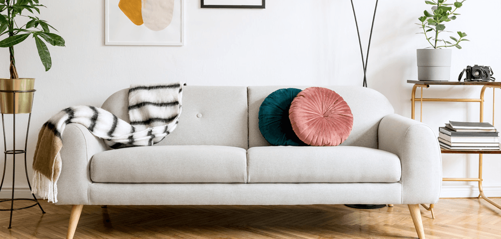 Modern light gray sofa with teal and pink pillows, zebra throw, potted plants, and minimalist decor in bright living room.