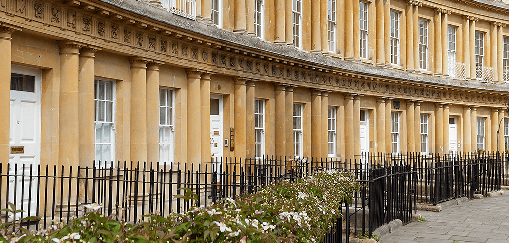 Elegant honey-colored Georgian townhouses with columns and tall windows behind black iron railings and flowering shrubs.