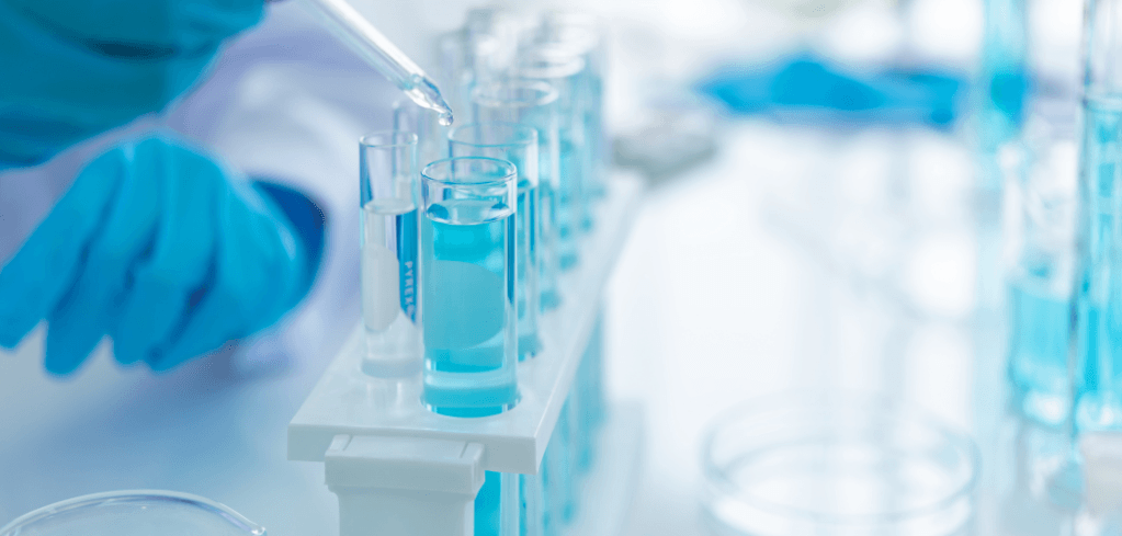 A gloved hand uses a pipette to add blue liquid into test tubes held in a white rack in a laboratory.