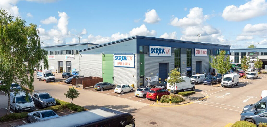 Aerial view of a Screwfix store in a retail park, open 7 days, with parked cars and vans under a blue sky.