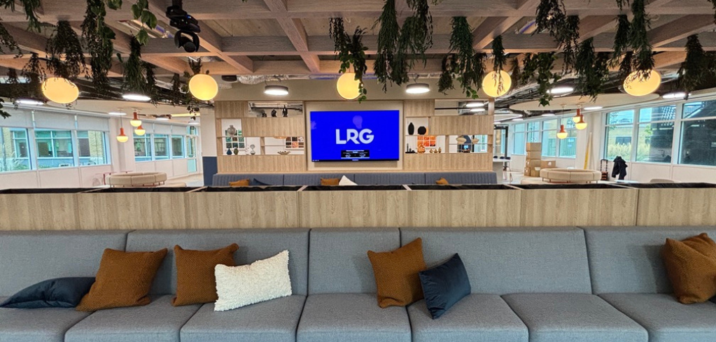 Modern office lounge with gray sofas, hanging plants, wooden accents, and a large blue screen displaying "LRG" logo.