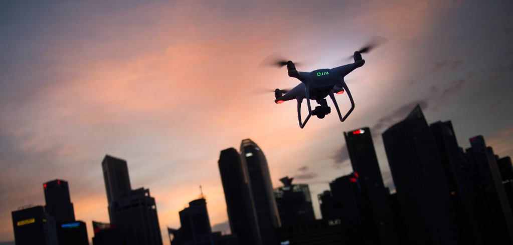 Drone flying over city skyline silhouette at sunset with orange and purple sky in background.