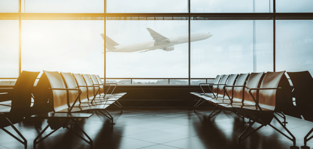 Empty airport gate seating area with large windows showing an airplane taking off in golden sunlight.