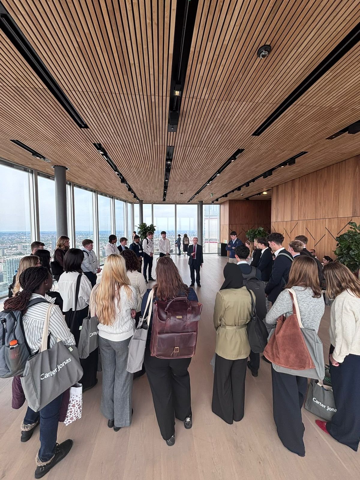 A group of young professionals with Carter Jonas bags gather in a modern high-rise room with panoramic city views and wood-paneled ceiling.