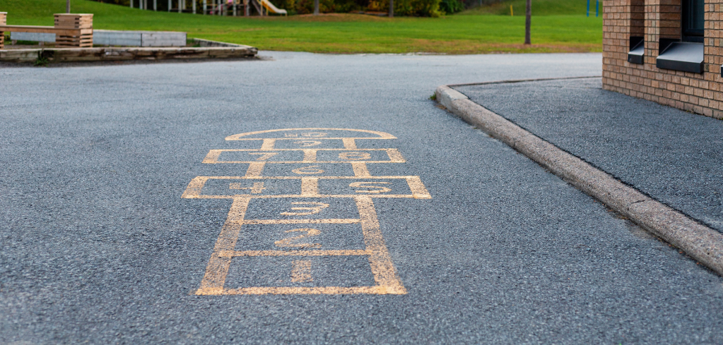Yellow hopscotch pattern painted on asphalt playground with green lawn and brick building in background.