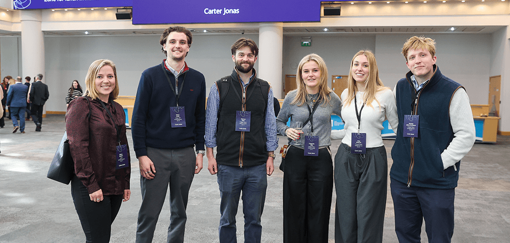 Group of six attendees wearing event badges standing together at a conference with "Carter Jonas" displayed on screen above.