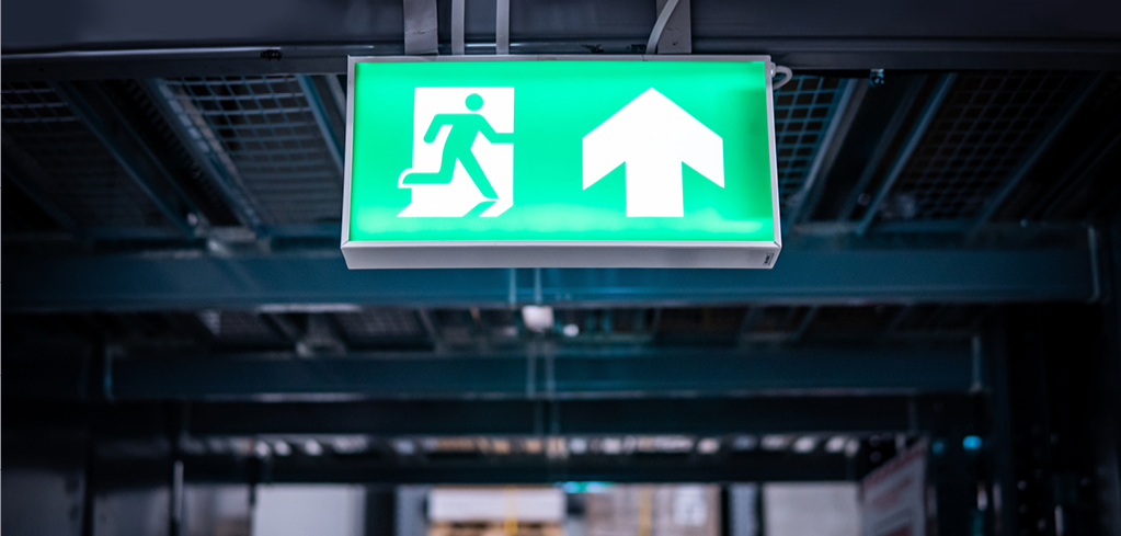 Green emergency exit sign with running figure and upward arrow mounted on dark industrial ceiling.