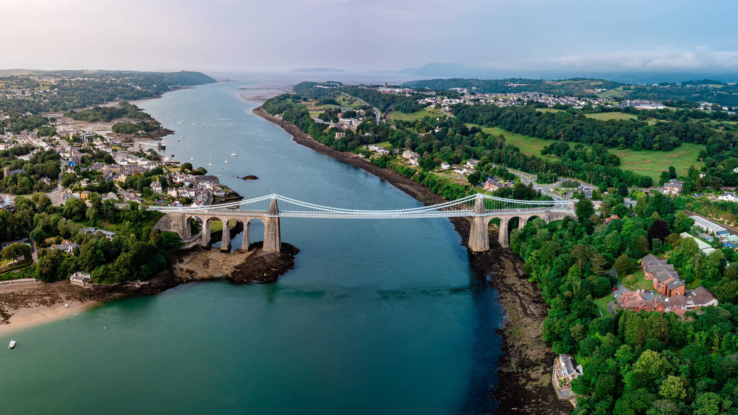 Aerial view of a suspension bridge spanning a blue river, surrounded by lush green trees and a small coastal town.