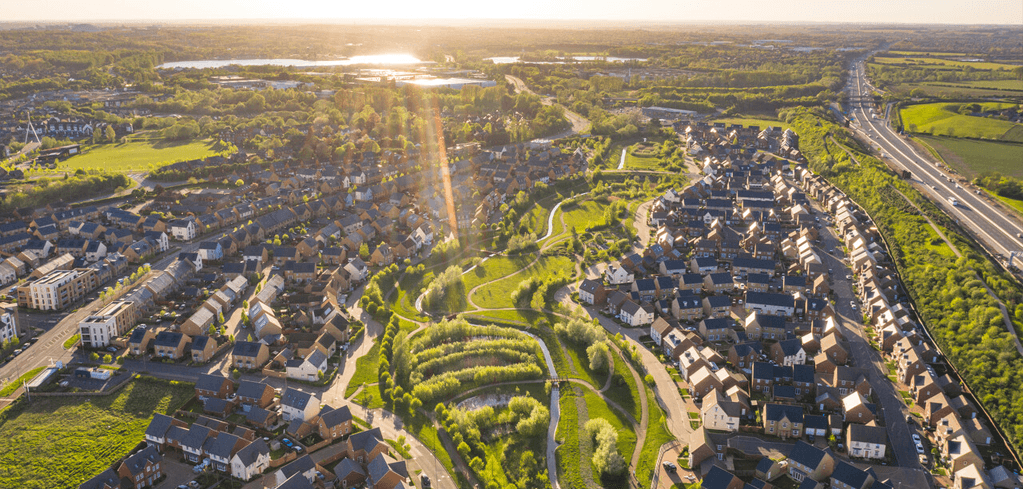 Aerial view of suburban housing development with central green space, lakes in distance, and sunset light illuminating the landscape.