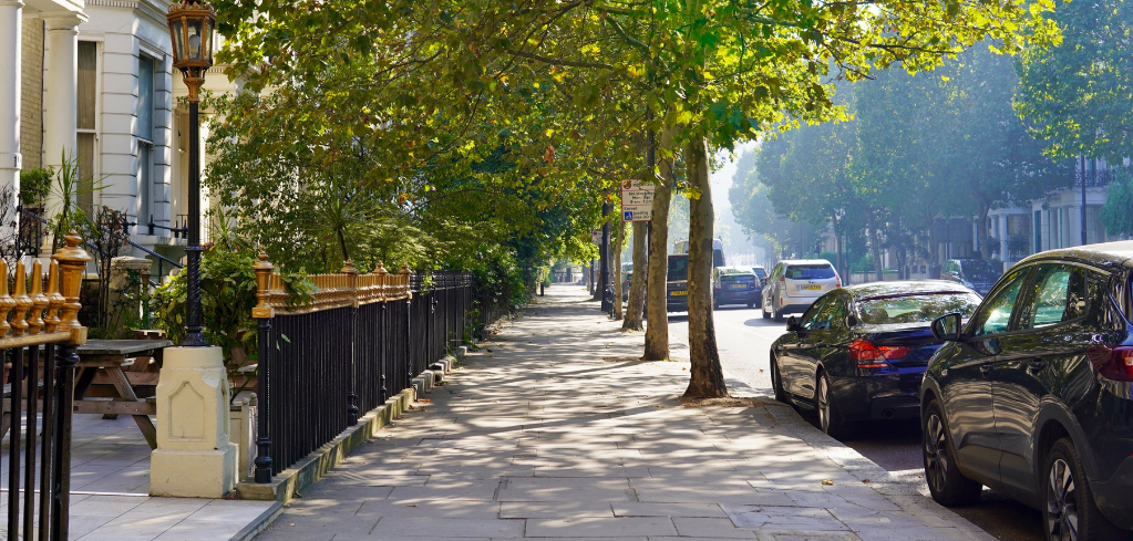 Tree-lined London street with elegant townhouses, wrought iron fences, and parked cars on a sunny morning.