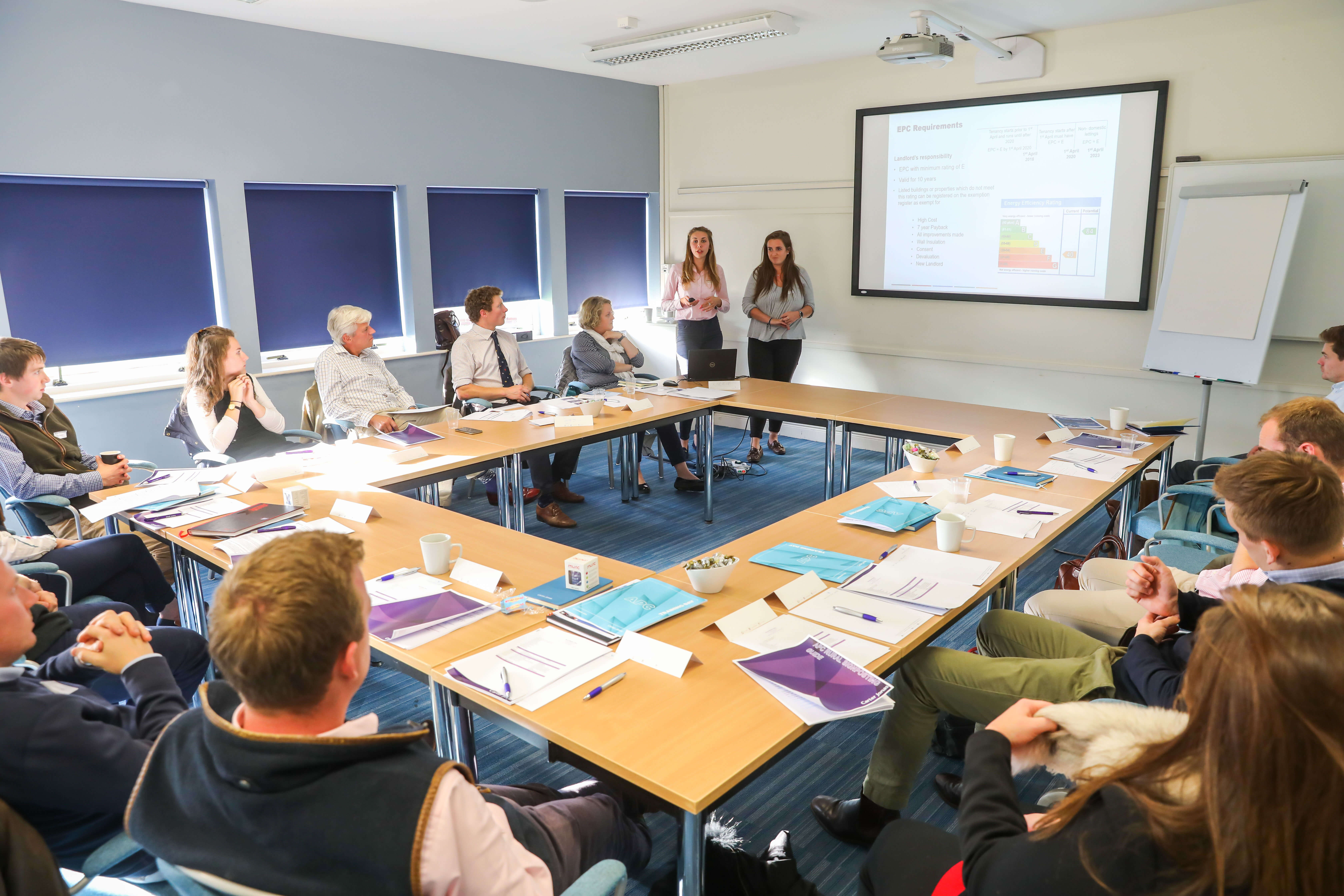 Two presenters standing by a projection screen showing EPC requirements to attendees seated around U-shaped conference table.
