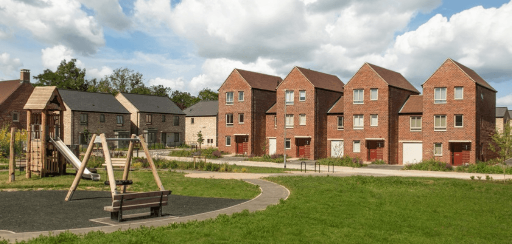 A residential neighborhood with red brick townhouses, a children's playground with a slide, green lawn, and a winding path.