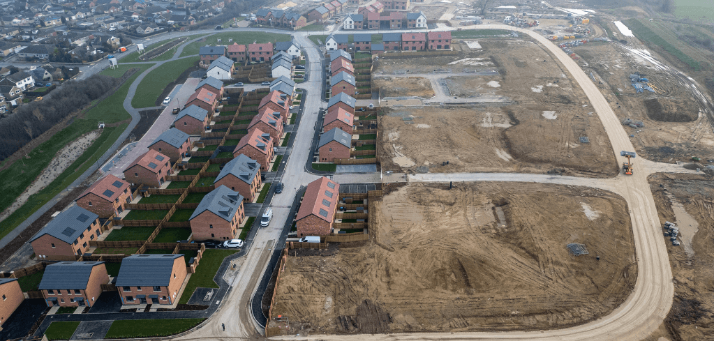 Aerial view of a new housing development with rows of brick homes alongside large areas of cleared land under construction.