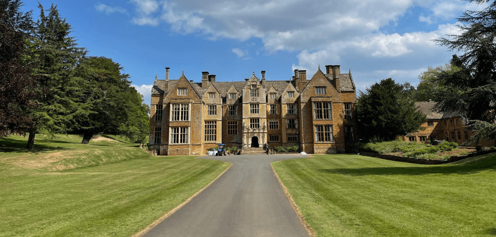 A grand English manor house with golden stone facade, flanked by tall trees and green lawns, under a blue sky with clouds.