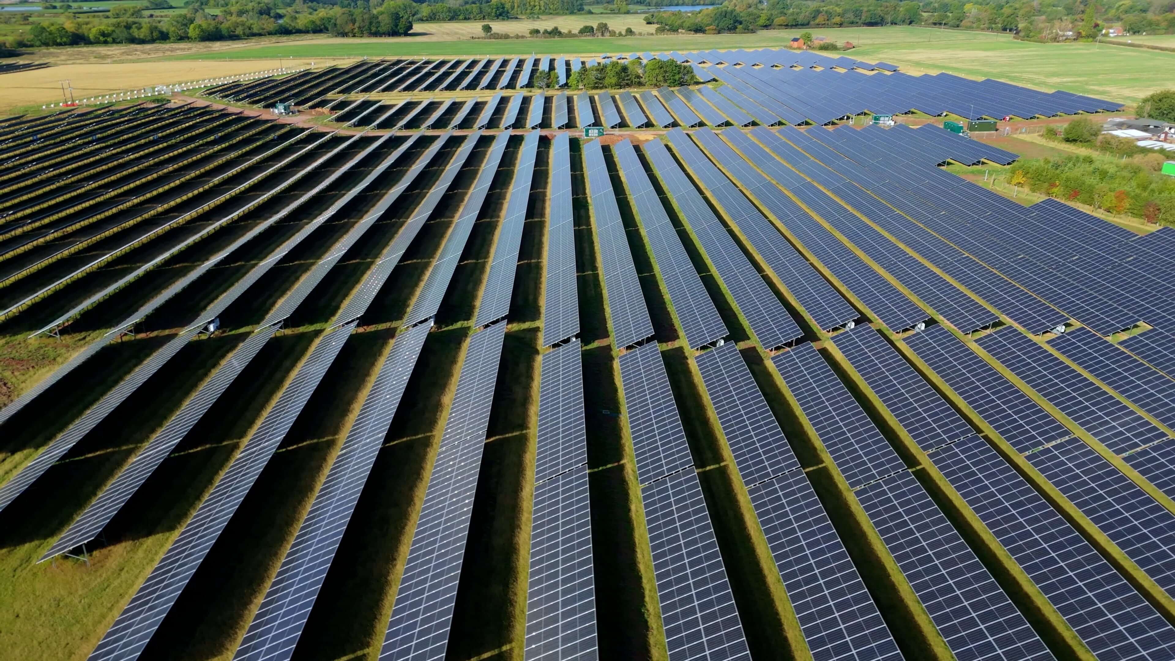 Aerial view of a large solar farm with rows of blue panels stretching across green countryside fields.