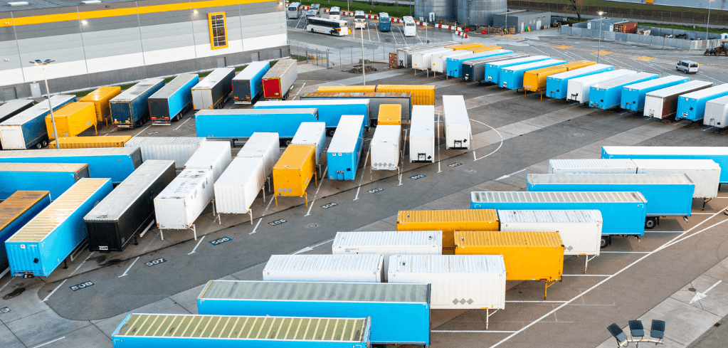Aerial view of a large logistics yard with rows of blue, yellow, and white truck trailers parked near a warehouse.