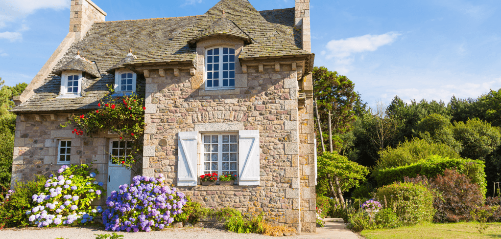 A charming stone cottage with blue shutters, colorful hydrangeas, and lush greenery under a bright blue sky.