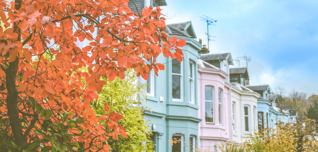 A row of colorful pastel townhouses beside a tree with vibrant red autumn leaves under a partly cloudy sky.