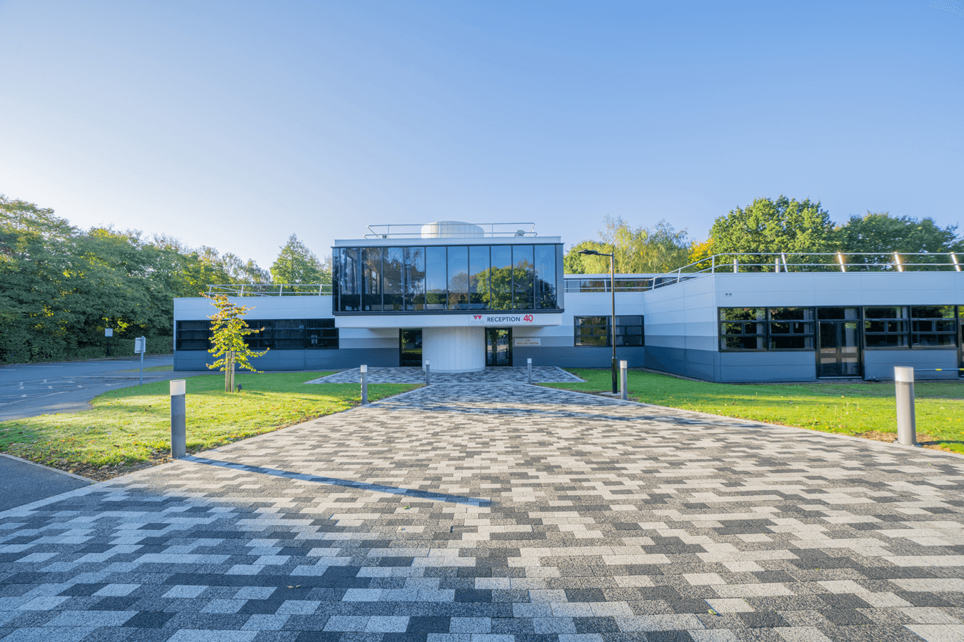 Modern office building with glass facade, paved entrance pathway, and surrounding greenery under clear blue sky.