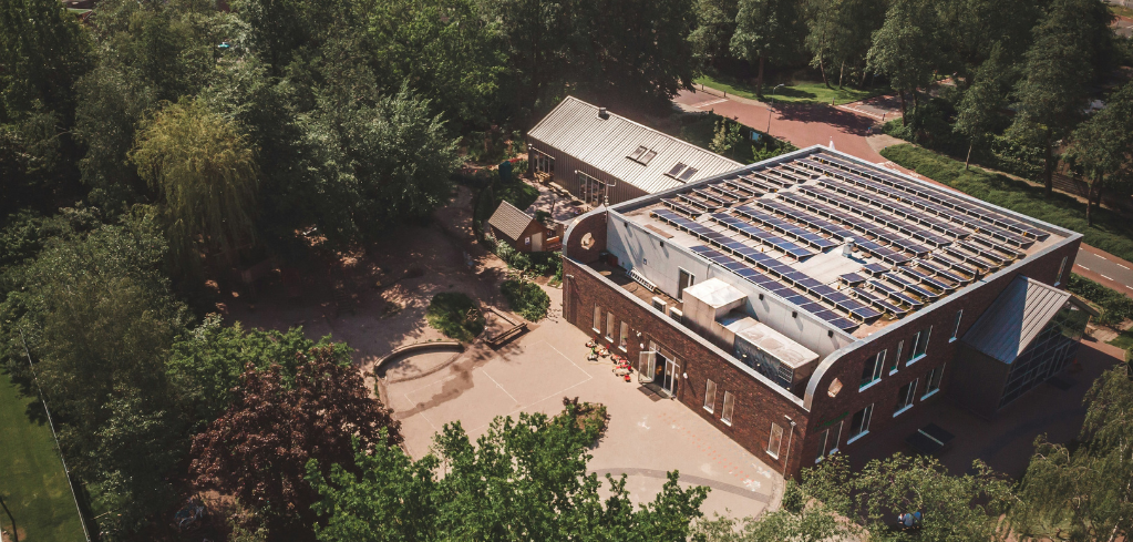 Aerial view of a brick building with solar panels on its flat roof, surrounded by lush green trees and a courtyard.