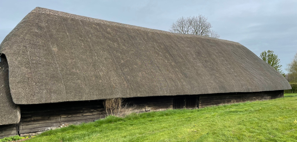 Traditional thatched-roof barn with wooden walls set on green grass under a cloudy sky.