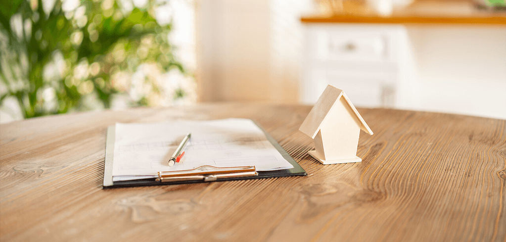 Small wooden house model next to an open notebook with pen on wooden table in bright, plant-decorated room.
