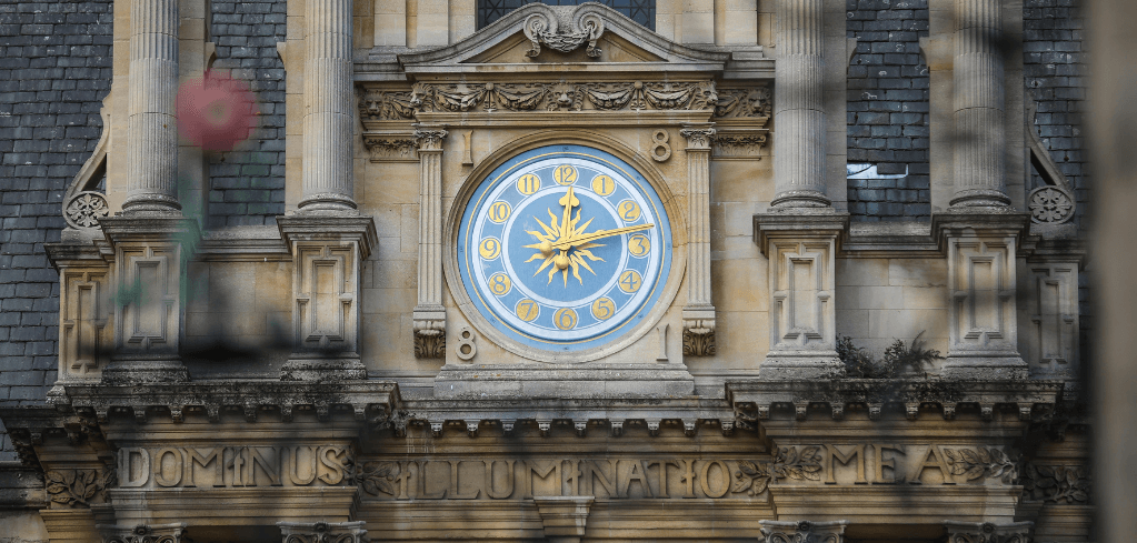 Ornate clock with gold sunburst design on blue face, set in stone facade of historic building with Latin inscription below.