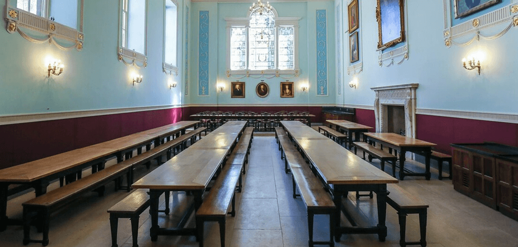 Elegant dining hall with light blue walls, burgundy wainscoting, stained glass windows, and long wooden tables with benches.
