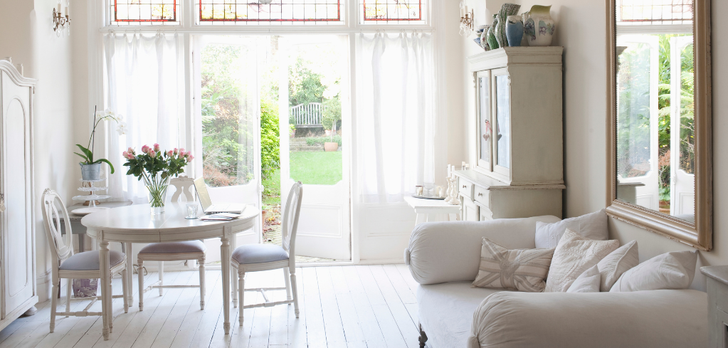 Bright white living room with dining table, sofa, and large windows overlooking a garden, creating an airy atmosphere.