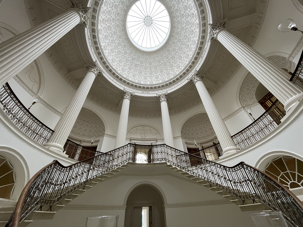 Elegant white domed rotunda with ornate ceiling, tall columns, and curved double staircase with wrought iron railings.