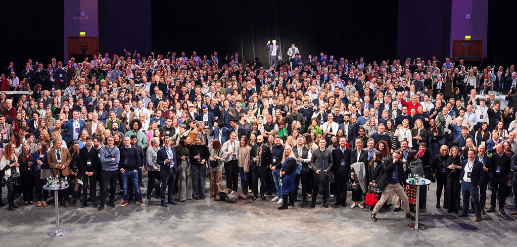 Large crowd of professionally dressed attendees gathered at a conference or convention in a dark-lit auditorium.