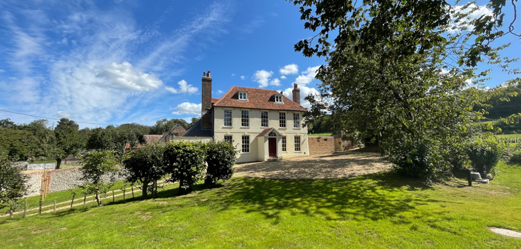 Traditional cream country house with red door and tiled roof surrounded by green lawn and trees under blue sky.