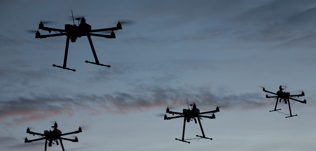 Four multi-rotor drones silhouetted against a blue-gray cloudy sky at dusk, flying in formation.