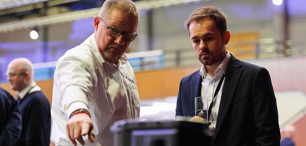 Two professionals examining equipment at a business event, one in chef whites pointing, one in navy suit observing.
