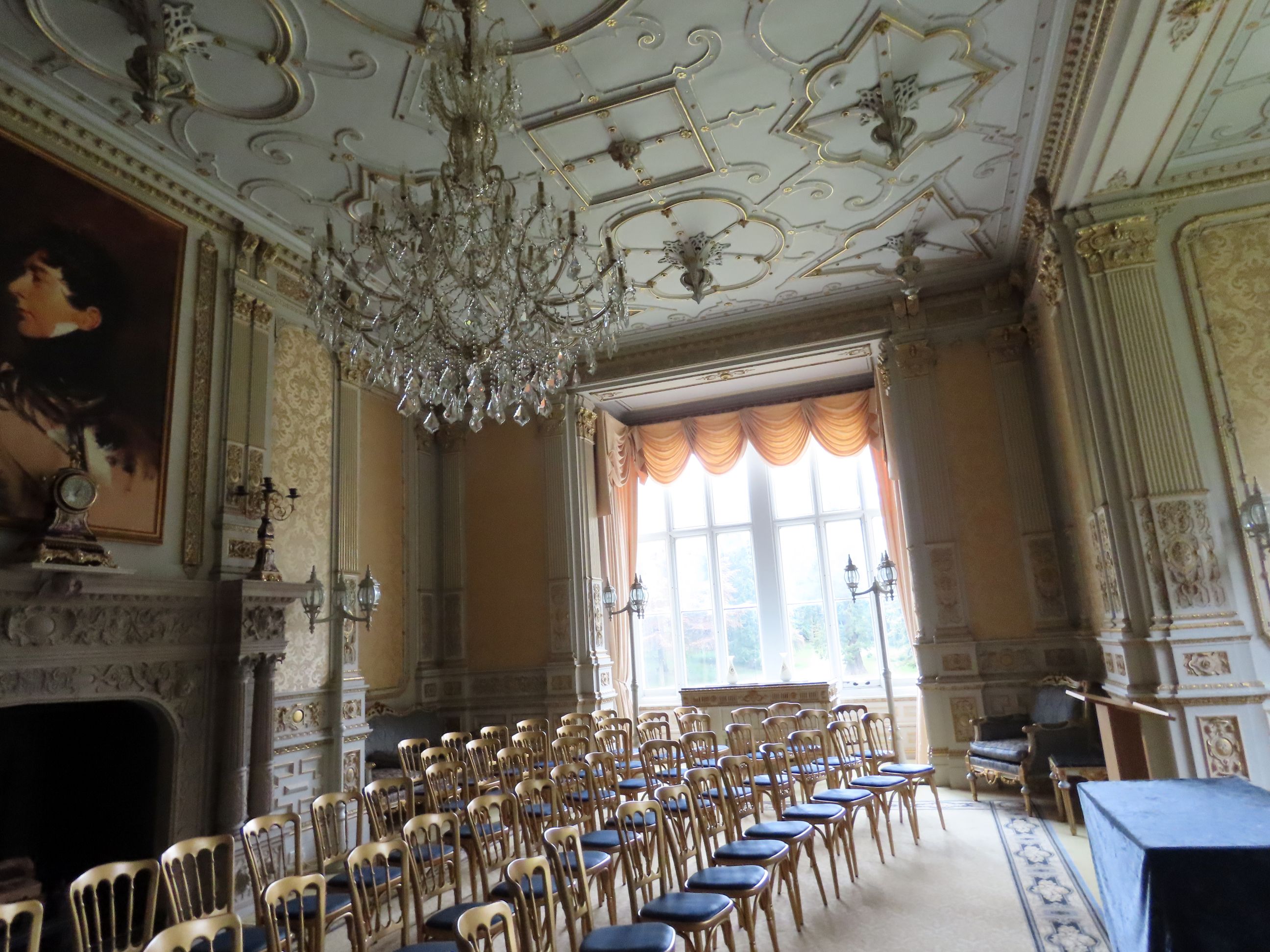 Ornate ballroom with gold chairs, crystal chandelier, decorative plaster ceiling, peach drapes, fireplace, and a portrait painting on the wall.