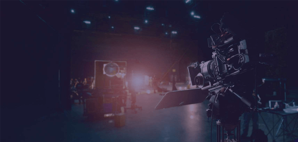 Professional video camera on a darkened film set with stage lights and equipment visible in the background.