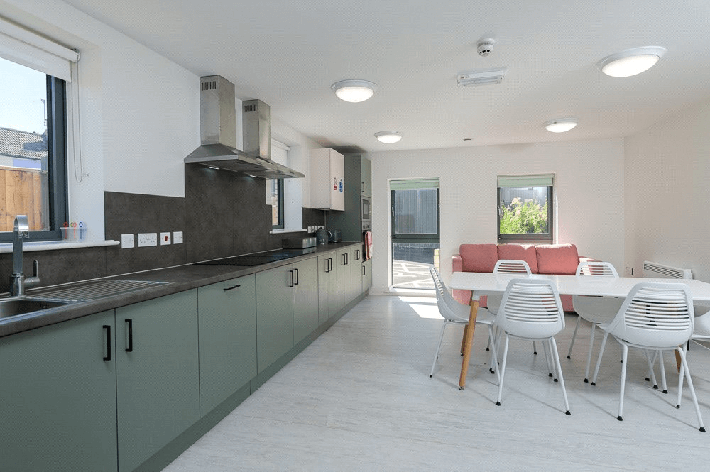 Modern kitchen with green cabinets, black countertops, and dining area featuring white table and chairs with pink sofa.