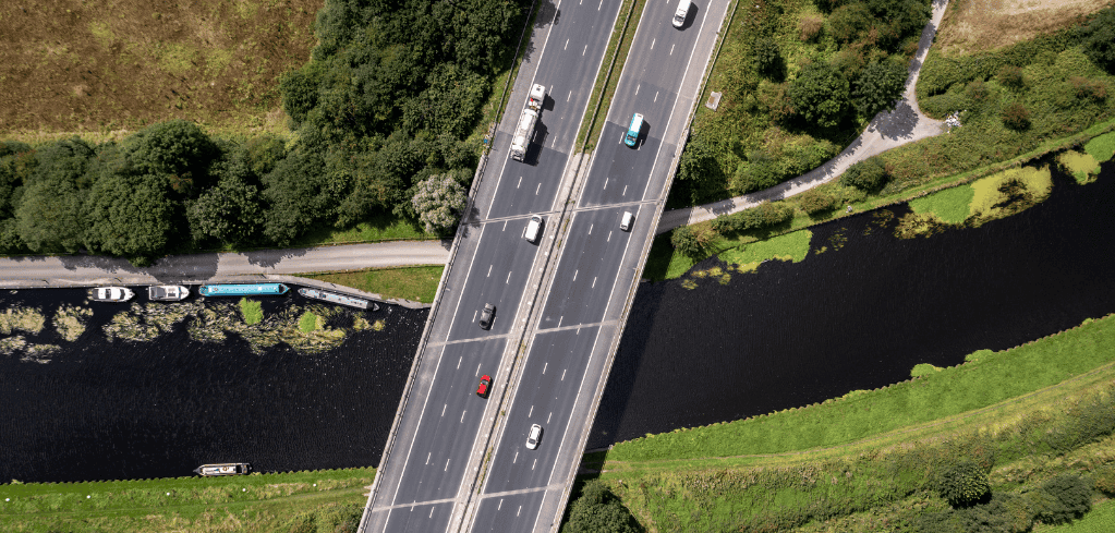 Aerial view of a highway bridge crossing a dark river, with cars traveling in both directions surrounded by green landscape.