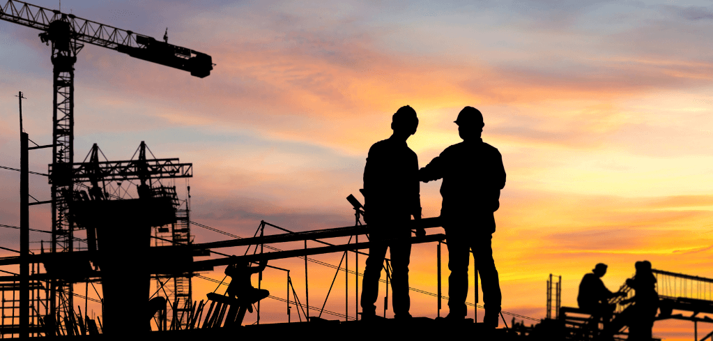 Silhouettes of construction workers on site at sunset, with a crane and scaffolding against an orange and purple sky.