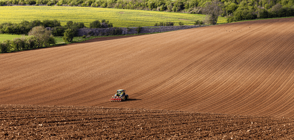 Tractor plowing a vast brown field, with green farmland and forest in the background under clear skies.