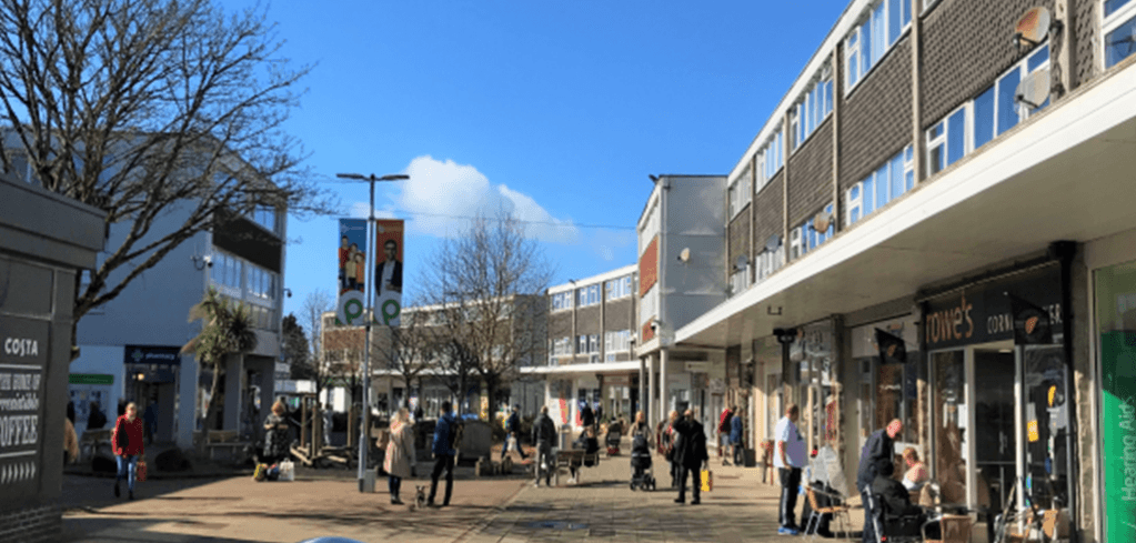 A busy pedestrian shopping street on a sunny day with shoppers, retail stores, bare trees, and a bright blue sky.