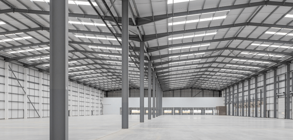 Interior of a large empty industrial warehouse with steel support columns, metal roof structure, and bright overhead lighting.