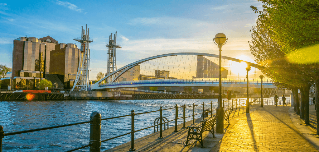 Sunny waterfront promenade with benches, a lamp post, trees, and a white arch bridge over a river with city buildings.