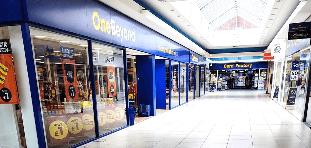 Interior shopping mall corridor featuring a blue OneBeyond store with price signs and a Card Factory shop in the background.