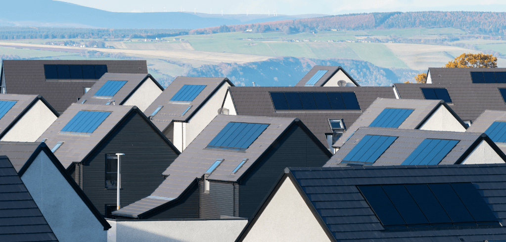 Rows of modern homes with dark rooftops fitted with blue solar panels, set against a scenic green valley and hills in Scotland, UK.