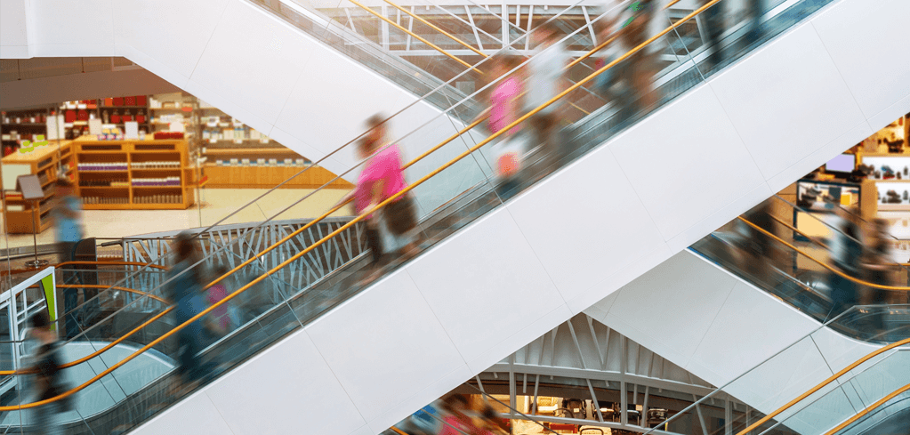 Blurred shoppers on multiple escalators in a modern shopping mall with white architectural features and retail stores.
