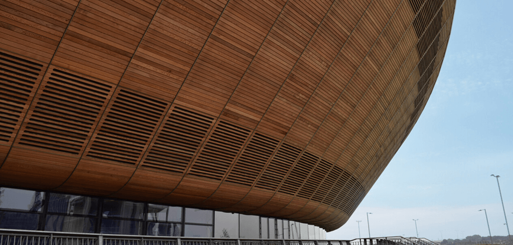 Modern curved wooden exterior of a stadium with glass walls beneath, against a blue sky.