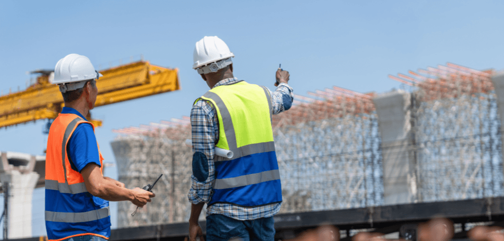 Two construction workers in white hard hats and safety vests survey a large building site with steel reinforcements and a crane.